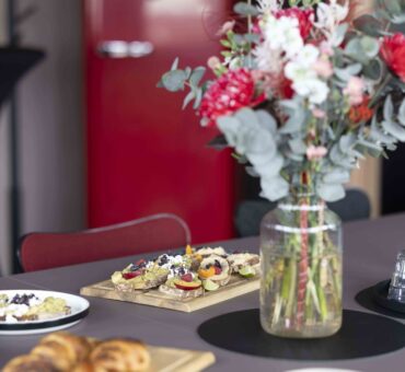 Frühstückstisch mit frischen Blumen in einer Glasvase, belegten Broten und Croissants – liebevoll gedeckt im MeetingRAUM Ingolstadt.