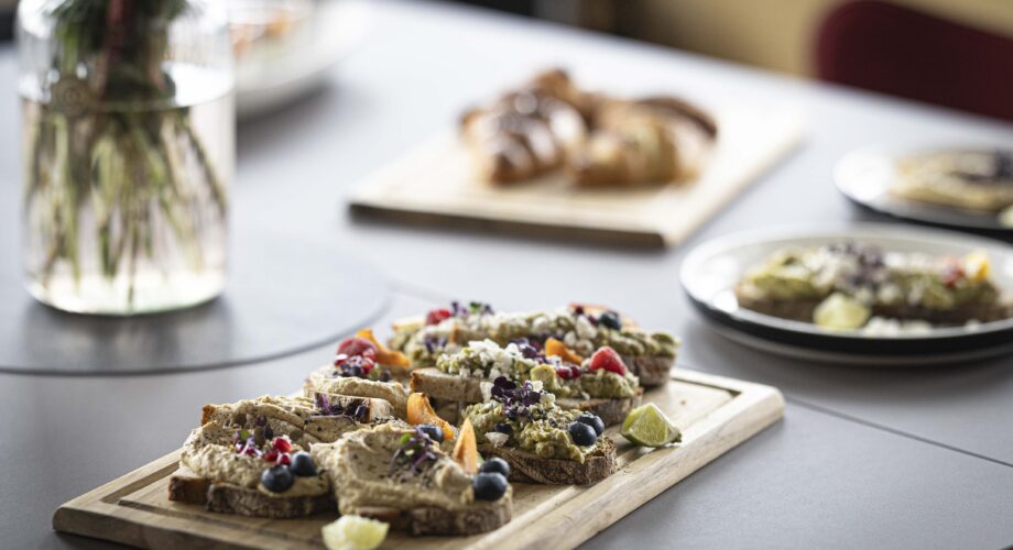 Frühstücks-Catering im MeetingRAUM Ingolstadt mit kreativ belegten Brotscheiben aus Hummus, Avocado, frischen Beeren und essbaren Blüten auf einem Holzbrett.