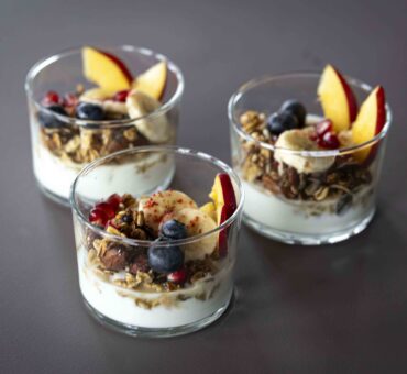 Drei Gläser mit Joghurt, Müsli und frischem Obst wie Blaubeeren, Banane, Granatapfelkernen und Pfirsich, serviert auf dunkler Tischplatte – Teil des Frühstücksangebots im MeetingRAUM Ingolstadt.