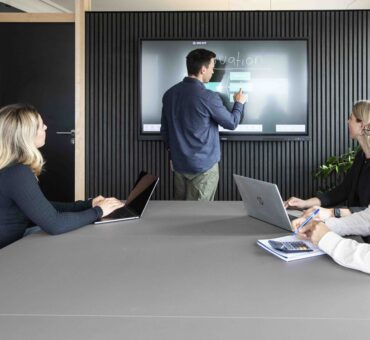 Ein Mann erklärt Inhalte an einem digitalen Touchscreen vor drei zuhörenden Personen mit Laptops – moderne Präsentation im MeetingRAUM Ingolstadt.