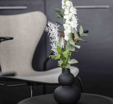 Schwarze Designvase mit weißen und grünen Trockenblumen auf einem Beistelltisch, im Hintergrund ein moderner Designerstuhl im MeetingRAUM Ingolstadt.