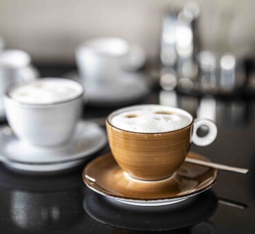 Frisch gebrühter Cappuccino in brauner Tasse steht unter dem Auslauf einer modernen Kaffeevollautomat im MeetingRAUM Ingolstadt. Im Hintergrund sind weitere Tassen und Untertassen zu sehen.