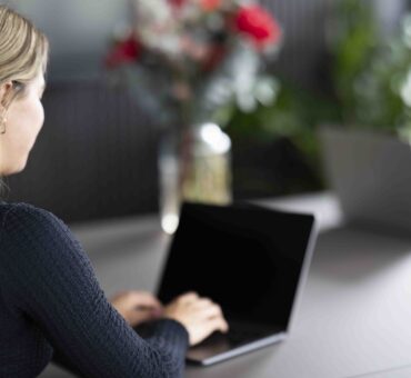 Eine Frau arbeitet lächelnd an einem Laptop in einem modernen, hellen Meetingraum in Ingolstadt. Im Hintergrund sitzen zwei weitere Personen mit Laptops und Notizblock, in einem professionellen Arbeitsumfeld mit Pflanzen und Tageslicht.