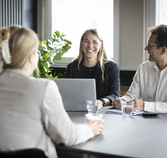 Vier Personen führen ein freundliches Business-Meeting an einem Tisch im MeetingRAUM Ingolstadt. Im Mittelpunkt sitzt eine lächelnde Frau mit Laptop, umgeben von Kolleg:innen in heller, moderner Umgebung mit Tageslicht und Pflanzen.